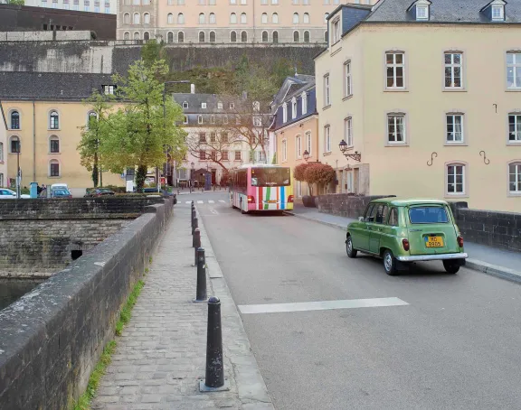A green car and a bus drive across a bridge with stone walls, lined with beige historical buildings, while the Alzette River flows below in Luxembourg City