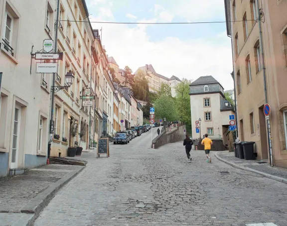 A steep cobblestone street in the old town of Luxembourg City, lined with historical buildings and parked cars, while two people run up the street