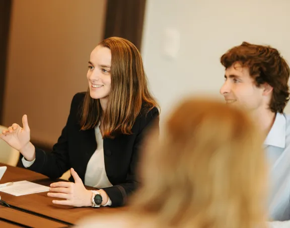 Two Stibbe lawyers, a woman and a man, sitting at a table in a meeting room. The woman is smiling, gesturing with her hand while speaking, and has a notepad in front of her. The man, also smiling, listens attentively. A blurred figure in the foreground faces them.