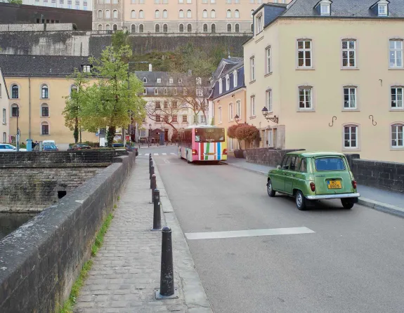 A green car and a bus drive across a bridge with stone walls, lined with beige historical buildings, while the Alzette River flows below in Luxembourg City