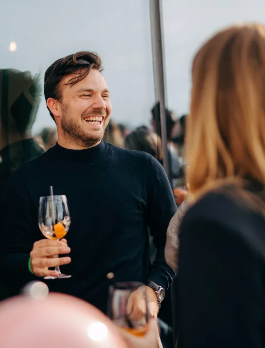 A man smiling broadly during a friendly conversation at a social event, holding a drink in his hand. The other people he is speaking with are facing away from the camera, engaged in the conversation with him.