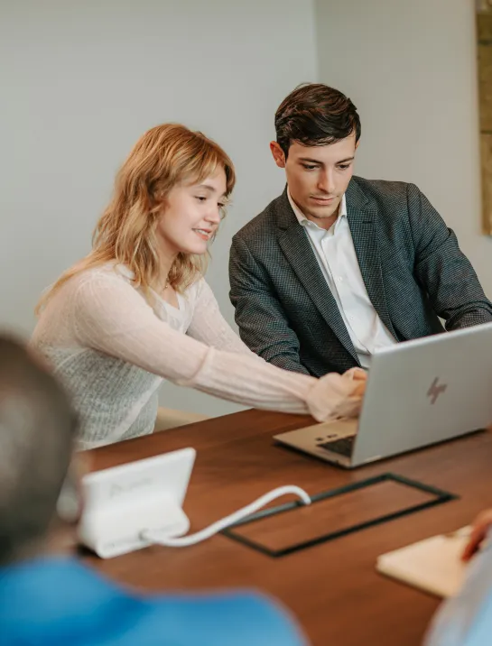 A woman sits at a desk next to a man, gesturing toward the screen of an open laptop as she explains something.