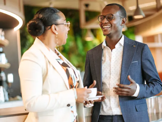 Two co-workers smiling warmly while engaging in a friendly conversation at the counter of the coffee bar at Stibbe in Brussels.