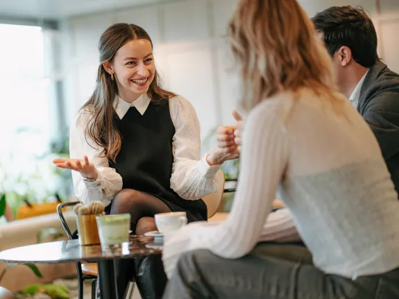 Two women and one man, all co-workers, engaging in a friendly conversation. The focus is on one woman, smiling broadly, while the other two are facing their backs to the camera, listening and smiling.