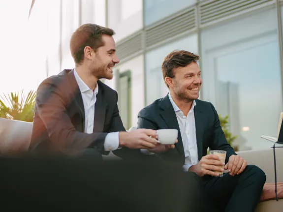 Two men seated side by side, smiling warmly as they engage with unseen conversation partners in front of them. 