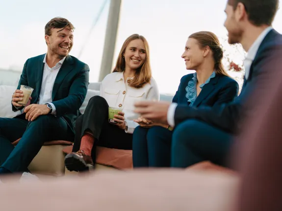 Group of four Stibbe co-workers, two women and two men, sitting on a comfortable outdoor sofa, enjoying drinks and engaging in friendly conversation.